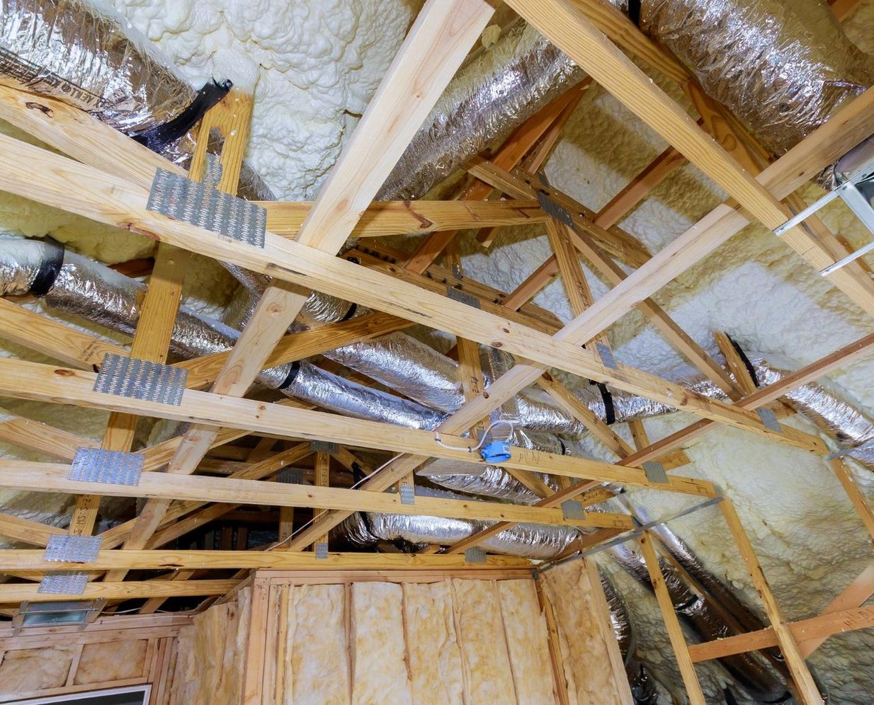 Spray foam insulation sealing the roof deck of an attic, with insulated ductwork running between the trusses